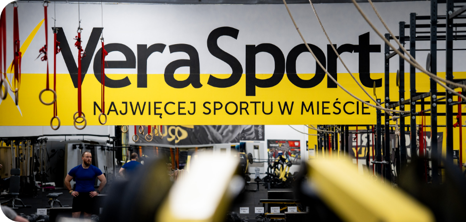 Strefa treningowa w Vera Sport z żółtym banerem ‘Najwięcej sportu w mieście’ i sprzętem do ćwiczeń.