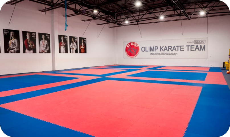 Sala treningowa Olimp Karate Team z matami w kolorze czerwonym i niebieskim oraz banerem na ścianie.